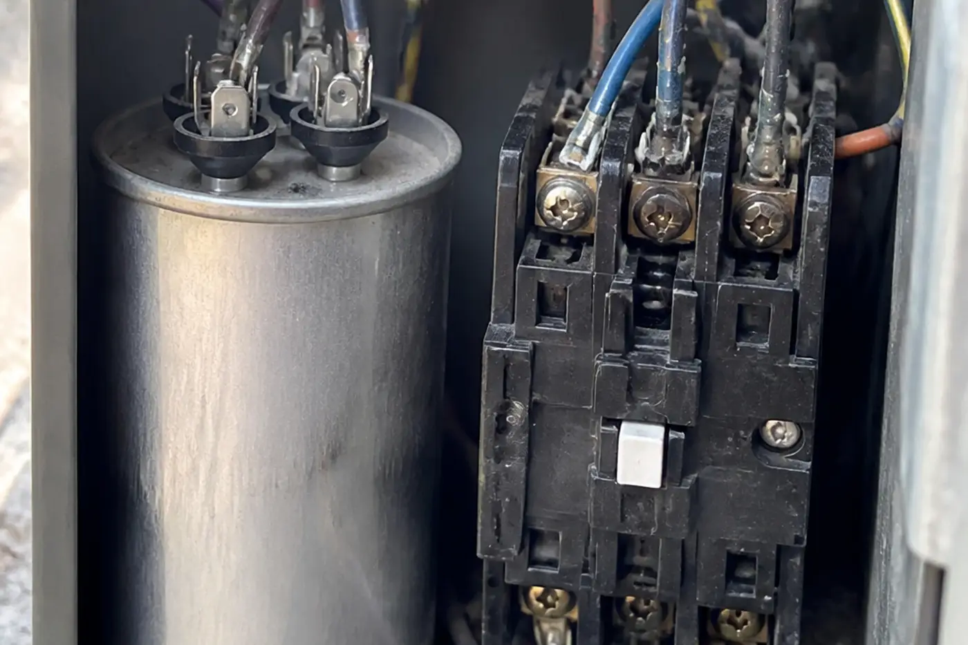 Round run capacitor next to a rectangular black AC contactor in an outdoor condenser electrical bay